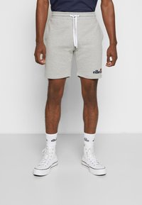 Shorts de survêtement gris clair avec une ceinture élastique et des cordons blancs. Présente le logo "ellesse" en bleu. Associés à des baskets montantes blanches.