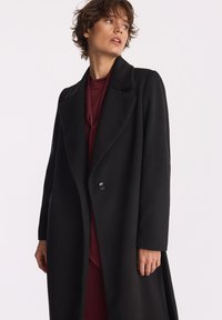 Kobieta z krótkimi włosami, ubrana w długi czarny płaszcz na burgundowej bluzce, patrzy w bok na tle jednolitego białego tła.