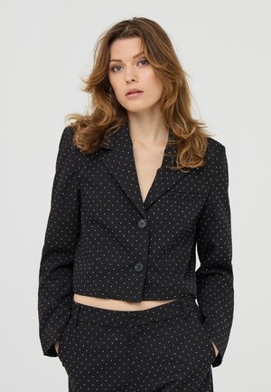 Donna con capelli castano chiaro mossi, che indossa un completo cropped nero con pois bianchi, mani in tasca e un'espressione facciale neutra.