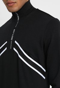 Detailní záběr osoby v černém topu s vysokým límcem, který má bílé diagonální pruhy a značkový zip. Důraz je kladen na límec a výstřih.