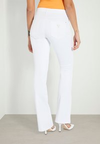 Jean évasé blanc avec taille haute, poches arrière brodées, porté avec des chaussures blanches à talons sur un sol gris clair.