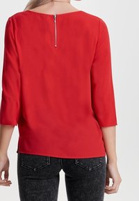 Blouse rouge à manches 3/4, texture lisse, encolure ronde et détail d'une fermeture éclair argentée au dos. Associée à un jean foncé.