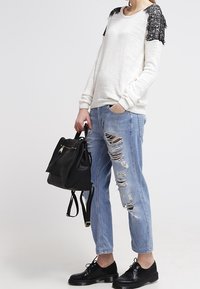 Personne portant un pull blanc avec des épaules en dentelle noire, un jean bleu clair déchiré, des chaussures noires à lacets et tenant un sac à dos en cuir noir.