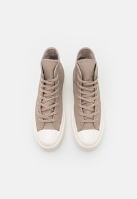 Wysokie trampki wykonane z jasnobrązowej skóry o teksturowanej powierzchni, z kremowym gumowym czubkiem i pasującymi sznurowadłami. Podeszwy są lekko podwyższone.