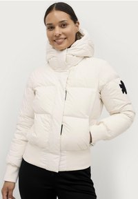 Giacca a vento in crema chiaro, con colletto alto, chiusura con zip e patch logo nero sulla manica. Realizzata in materiale liscio e isolante.