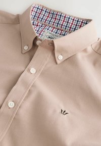 Bežová košile s límečkem z bavlněné směsi s bílými knoflíky. Uvnitř má červenobílo-modrý kostkovaný vzor. Vyšité logo na přední straně.