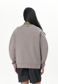 Šedá oversized mikina s žebrovanými manžetami a spodním lemem, s padlými rameny a smyčkou na zadní straně, nošená přes černé kraťasy.