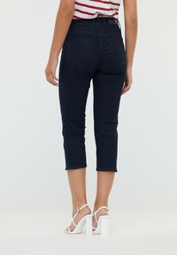 Jean cropped bleu foncé avec un design ajusté et des poches arrière, associés à des sandales à talons blocs blanches. Fond uni.