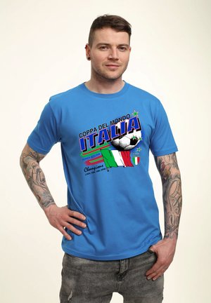 Mężczyzna z tatuażami noszący niebieską koszulkę piłkarską "Coppa del Mondo Italia" z włoską flagą i latami mistrzostw, stojący z ręką na biodrze.