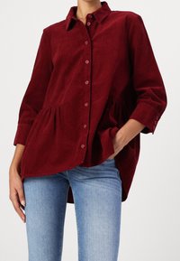 Bordeaux corduroy overhemd met een gestructureerde kraag, knoopsluiting aan de voorkant en geplooide peplumdetail; gecombineerd met blauwe jeans.