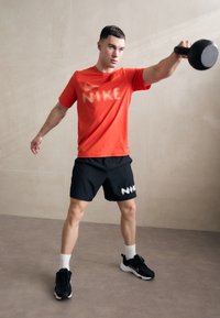 Punane Nike T-särk stiliseeritud logoga, mustad lühikesed püksid valgete graafikatega ja mustad sportlikud jalanõud; kettlebell hoitakse üle pea.