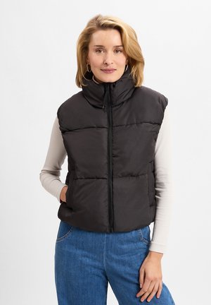 Svart puffer vest med høy krage og glidelås foran, med en quiltet design og to sidelommer. Båret over en lys langermet topp.