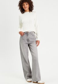 Pull en tricot blanc avec des motifs en losange texturés, associé à un jean gris ample taille haute. Le modèle porte des chaussures décontractées à enfiler.