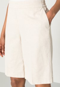 Světlé béžové šortky vyrobené z texturované tkaniny, s elastickým pasem a bočními kapsami. Střih je volný a šortky dosahují ke kolenům.