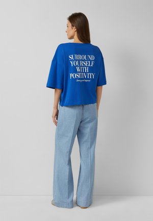 Donna con lunghi capelli castani che indossa una maglietta blu larga con la scritta "Surround yourself with positivity" e jeans chiari a gamba larga, in piedi di lato.