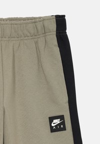 Artiškas žiūrėjimas į smėlio spalvos ir juodas Nike Air treningo kelnes su elastine juostele ir Nike Air logotipo etikete kairėje pusėje.