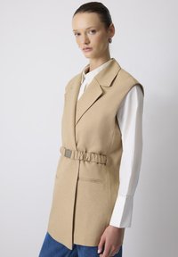 Beige mouwloze blazer met een taille die is getailleerd, kraag en zijzakken, gelaagd over een witte blouse, met een gladde, gestructureerde afwerking.