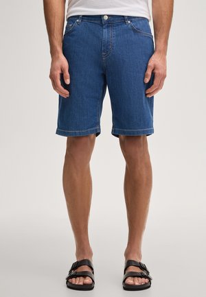 Mann trägt blaue Jeansshorts, weißes Hemd und schwarze Sandalen mit Schnalle, steht vor einem einfarbigen hellen Hintergrund.