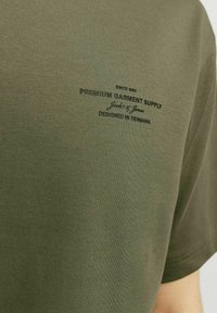 Zelena pamučna majica s okruglim izrezom i kratkim rukavima. Ispis teksta u crnoj boji: "Premium Garment Supply" i "Dizajnirano u Danskoj."