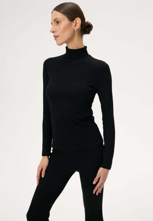 Kobieta o brązowych włosach związanych w niski kok, nosząca srebrne kolczyki, obcisły czarny golf i czarne spodnie, pozująca z rękami na biodrach na białym tle.