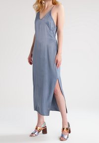 Vestido de satén gris con escote en V y tirantes finos, con una abertura lateral. Combinado con sandalias de tacón cuadrado en colores metálicos.