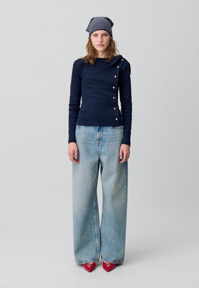 Maglietta a maniche lunghe navy con bottoni dorati, abbinata a jeans a gamba larga blu chiaro e tacchi a punta rossi. Cappello a righe navy e bianche.