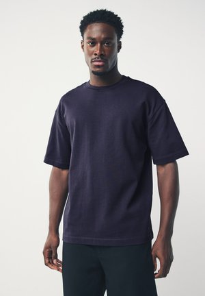 Man staat frontaal, draagt een effen donkerblauw oversized T-shirt met korte mouwen en zwarte broek tegen een lichte achtergrond.