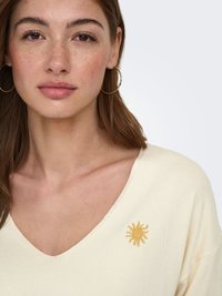Jeune femme avec de légères taches de rousseur, portant des boucles d’oreilles créoles dorées et un haut crème à col en V avec un petit soleil jaune brodé près de la clavicule.