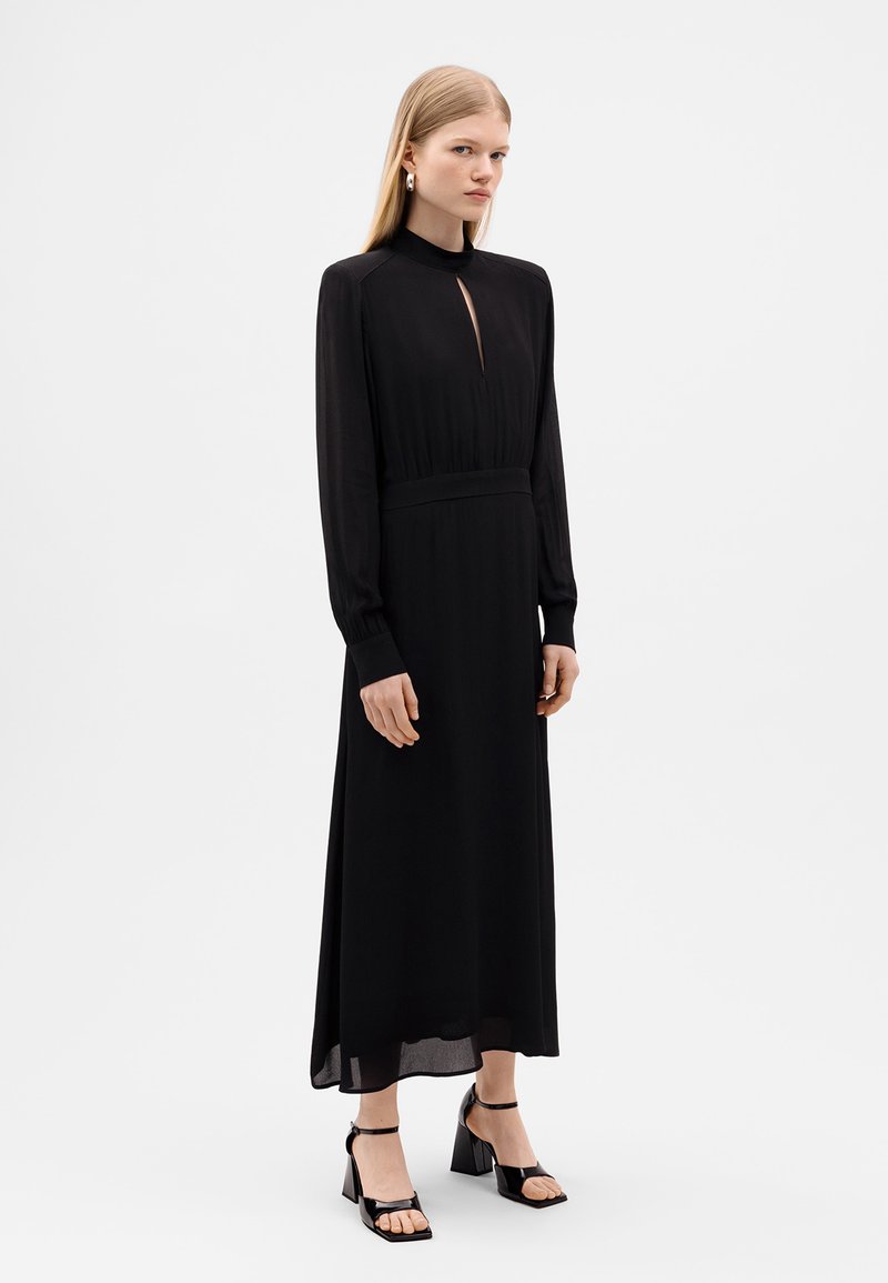 Czarna sukienka z długim rękawem, z wysokim kołnierzem i dekoltem w kształcie łezki z przodu, dopasowana w talii i z luźnym dołem. Połączona z czarnymi sandałami na obcasie.