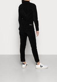 Jupe-pantalon en velours côtelé noir au design ajusté, avec des manches longues et des détails de boutons. Des sneakers blanches avec un logo noir complètent la tenue.