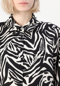 Crno-bijela košulja s uzorkom zebra pruga, s ovratnikom i crnim gumbima sprijeda. Glatka tekstura tkanine.