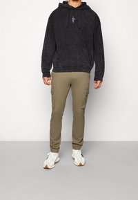 Sort fleecetrøje med hætte, frontlomme og lilla grafisk detalje, kombineret med olivengrønne cargobukser med sidelommer og elastiske manchetter.