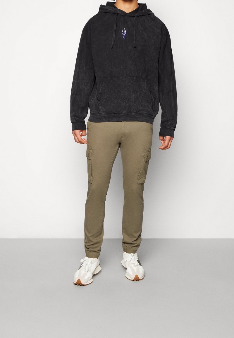 Sort fleecetrøje med hætte, frontlomme og lilla grafisk detalje, kombineret med olivengrønne cargobukser med sidelommer og elastiske manchetter.