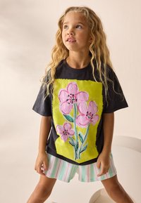 Juodas marškinėlis su ryškia žalia gėlių raštu, kuriame yra rožinių gėlių ir žalių lapų, derinamas su šviesiais dryžuotais šortais.