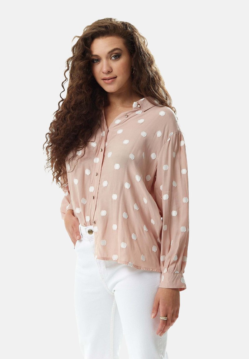 Light pink button-up shirt with white polka dots, featuring a relaxed fit, long sleeves, and a slightly curved hem. Paired with white jeans.