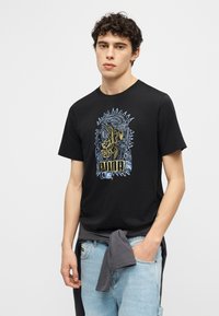 Νεαρός με σγουρά μαλλιά, φορώντας μαύρο μπλουζάκι Puma με σχέδιο και ανοιχτόχρωμο τζιν, με σκούρο μπουφάν δεμένο γύρω από τη μέση, στέκεται μπροστά σε λευκό φόντο.