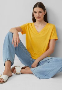 T-shirt jaune à manches courtes en V, fabriqué en tissu doux, associé à un jean large bleu clair et des sandales blanches avec un détail de boucle.