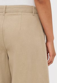 Beige laiad sääred, millel on tekstureeritud kangas, vööd, ja küljetaskud, nähtavad tagant, käsi osaliselt näha.
