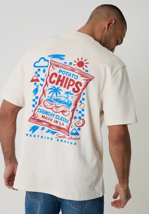 Uomo con maglietta bianca con una grafica colorata di una borsa di patatine e palme, con la scritta "Crunchy Classic Made in LA, Santa Monica."