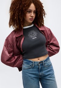 Νέα γυναίκα με σγουρά μαλλιά, ντυμένη με μπουφάν βουργουνδίας, μαύρο κοντό μπλουζάκι με γραφικό ήλιο και μπλε τζιν ψηλόμεσο, με τα χέρια στη μέση.