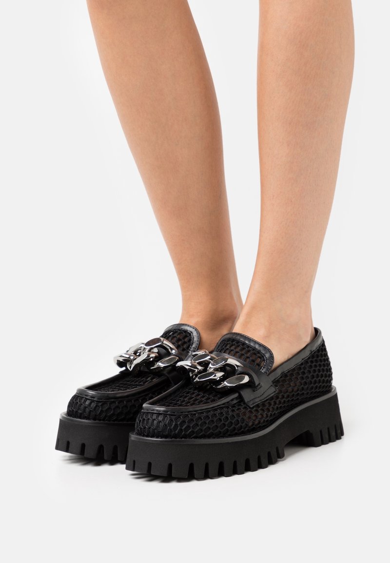 Mocassins en mesh noir avec semelles épaisses à crampons et grand détail chaîne argentée portés pieds nus.
