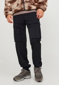 Svarta cargobyxor med flera fickor, elastiska muddar och en slät textur. Bärs med grå sneakers och en mönstrad brun fleecejacka.