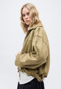 Νεαρή γυναίκα με κυματιστά ξανθά μαλλιά που φοράει μια φαρδιά ανοιχτόκαφε σουέντ ζακέτα πάνω από λευκό πουκάμισο και μαύρο παντελόνι, μπροστά σε απλό φόντο.