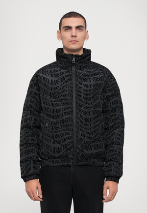Black zip-up jacket featuring a textured pattern of bold, repeating text. High collar and elasticized cuffs. Lightweight design.