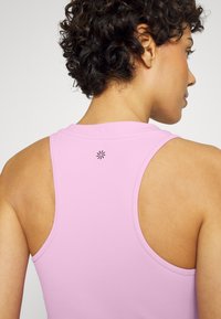 Top desportivo rosa claro com decote redondo e aberturas largas para os braços. Apresenta um pequeno logótipo preto nas costas, textura de tecido suave.
