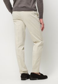Beige Baumwollhose mit Slim-Fit, seitlichen Taschen und einer Gesäßtasche. Trägt dazu dunkelbraune Wildleder-Schlüpfschuhe, von hinten betrachtet.