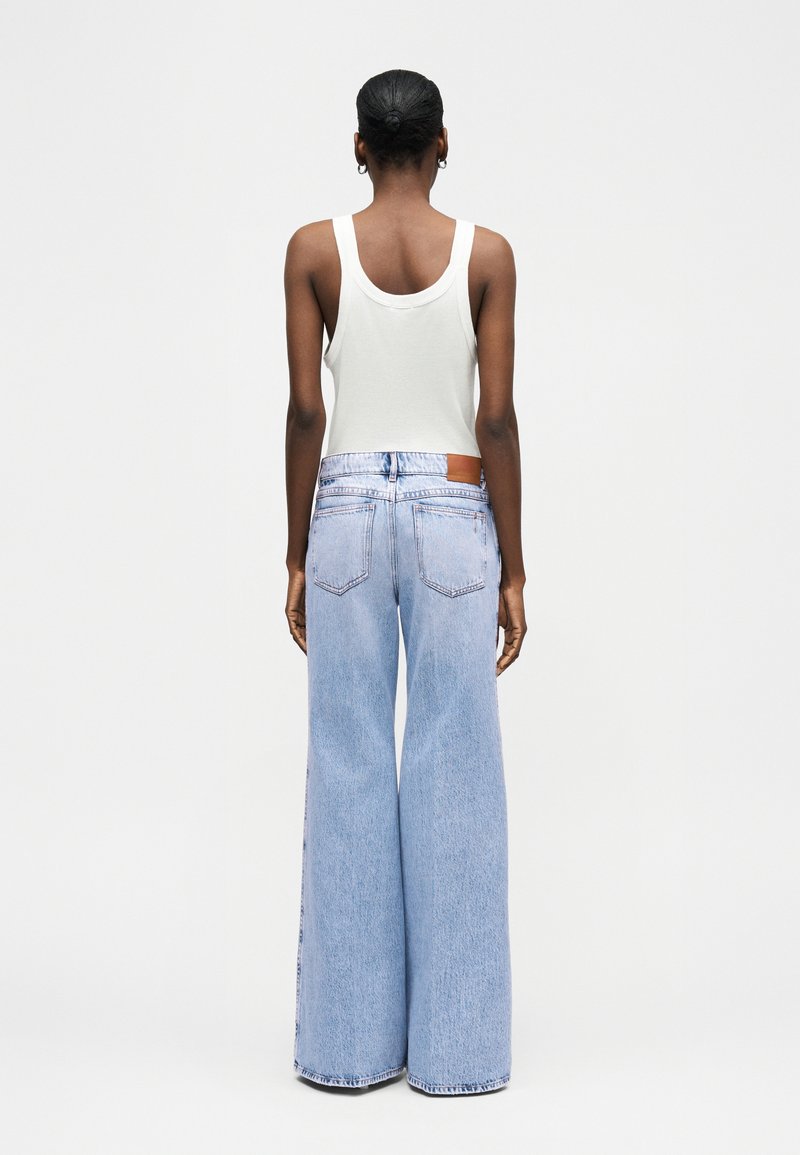 Vrouw met kort haar, gekleed in een witte tanktop en lichtblauwe wijde jeans met hoge taille, van achteren gezien tegen een effen achtergrond.