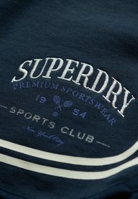 Navy katoenen sweatshirt met witte geborduurde tekst "SUPERDRY", sportclublogo en twee witte strepen aan de onderkant. Geweven stof.