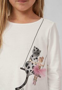 Camicia bianca a maniche lunghe con una grafica di una leopardessa e una ragazza su un'altalena. La leopardessa ha dei motivi a forma di cuore neri; la ragazza indossa un tutù rosa.