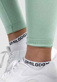 Lichtgroene legging met een gladde textuur, met een subtiele logo bij de enkel. Gecombineerd met witte sneakers en zwart-witte sokken.